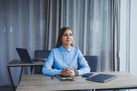 A beautiful woman in glasses sits at a table in the office and works at a laptop. Corporate work in the office. Modern woman workplace manager in classic clothes