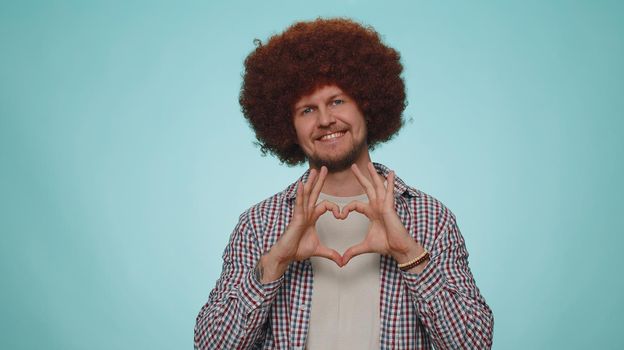 Man in love. Smiling man 30s with lush hairstyle makes heart gesture demonstrates love sign expresses good feelings and sympathy. Young adult guy boy isolated alone on blue studio wall background