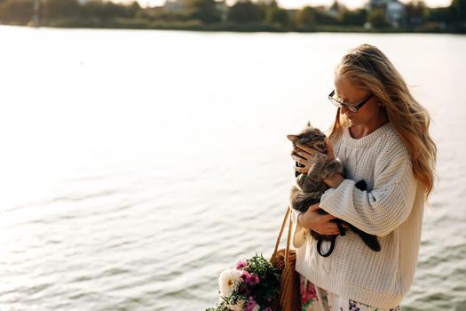 Young blonde, girl, woman with glasses holds a cat breed Scottish Straight, the cat bites. Walk with pet near the lake, river at sunset on the beach in Spring