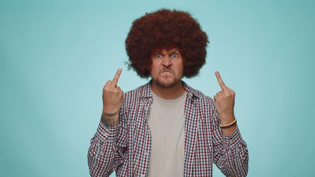 Aggressive crazy hooligan man showing around his middle finger, demonstrating protest with impolite rude gesture of disrespect, rejecting communication. Young adult guy isolated on blue background