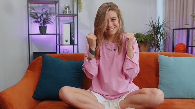 Happy caucasian girl shouting, celebrating success, winning, goal achievement, good news, raising fists in gesture I did it, lottery luck at home apartment. Young woman indoors in living room on couch