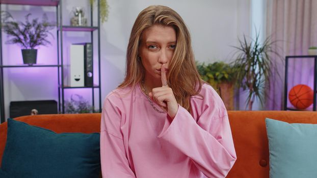Shh be quiet please. Adult caucasian girl presses index finger to lips makes silence hush gesture sign do not tells gossip secret. Young woman at home apartment living room sitting on orange couch