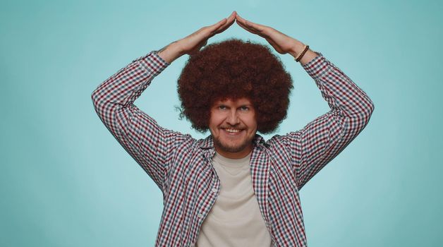 Happy bearded man playing childish catching up game, feeling in safe making roof above head with hands, insurance, security service. Young adult guy boy isolated alone on blue studio background