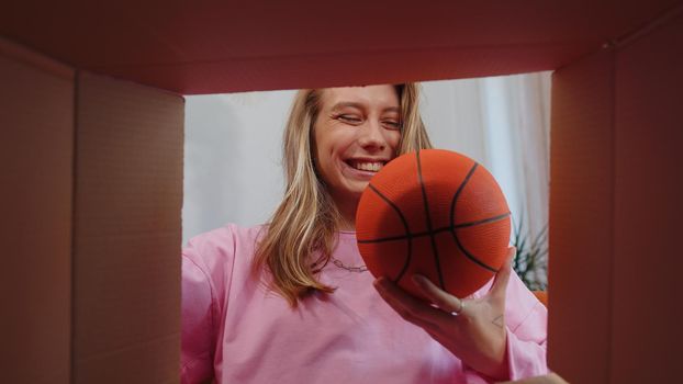 Happy surprised winner girl unpacking delivery parcel at home. Smiling satisfied young woman shopper, online shop customer opening cardboard box receiving purchase gift ball by fast postal shipping