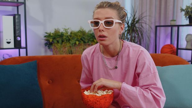 Excited adult girl sitting on sofa eating popcorn and watching interesting tv serial, sport game, film, online social media movie content online at home. Young woman enjoying domestic entertainment