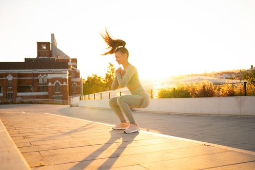Sporty woman doing jumping squats exercises on stairs in park. Bodyweight training on sunset. Young adult fitness woman exercising outdoor. European woman squats. Athletic caucasian female workout