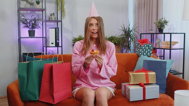 Happy adult girl wears festive birthday hat hold cupcake makes wish joyful congratulating blowing burning candle on cake. Young woman celebrating anniversary party at home room apartment on couch