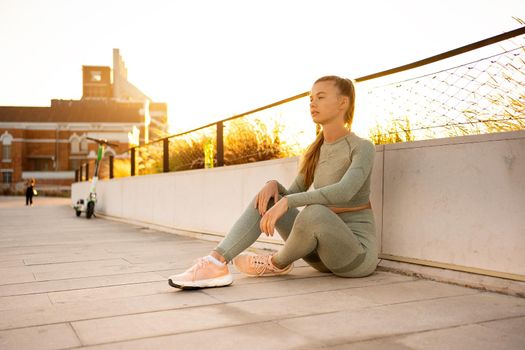 Sportswoman sitting on concrete floor in summer city after morning workout looking away confident. Active lifestyle. Fit caucasian female in sportswear ready for jogging. Fitness woman on sunset