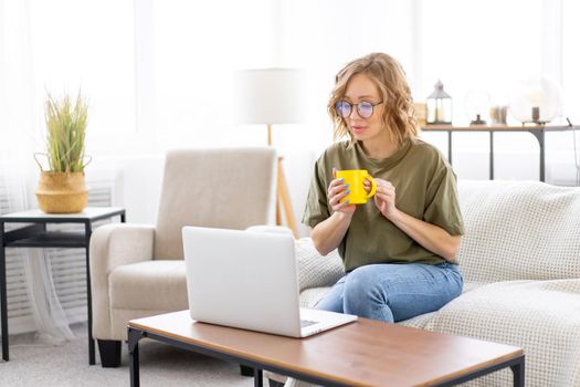Woman with glasses use laptop typing keyboard sitting couch big window background home interior Freelance female working from home Distance learning student relaxing watch lessons video conference.