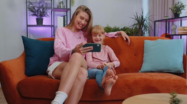 Little small child kid daughter having fun with mother or nanny enjoying using smartphone, showing funny face mask app on modern gadget cellphone looking at mobile screen laughing sit at home couch