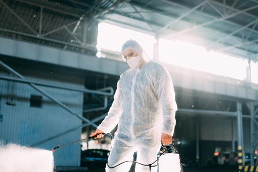 Man dressed white protective overalls spraying surface antibacterial sanitizer sprayer during quarantine Caucasian person protective suit disinfects parking during covid pandemic backlight