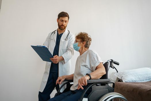 Doctor in mask explaining to a sick patient in wheelchair details of treatment. High quality photo