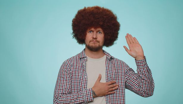 I swear to be honest. Sincere responsible man raising hand to take oath, promising to be honest and to tell truth, keeping hand on chest. Young adult guy boy isolated alone on blue studio background