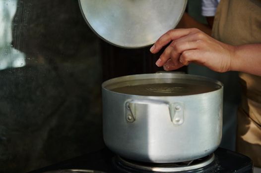 Close-up. Cropped view of a housewife standing by a kitchen table with electric stove, opening the lid of a metal saucepan, dipping raw dumplings into a boiling water, brewing Ukrainian vareniki.