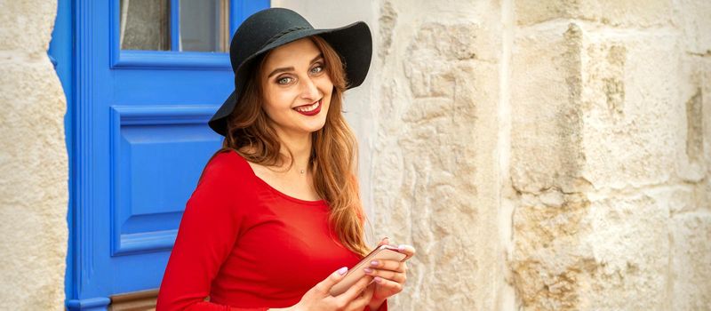 Portrait of tourist young caucasian woman in a red long dress and black hat with suitcase sitting on the stairs outdoors