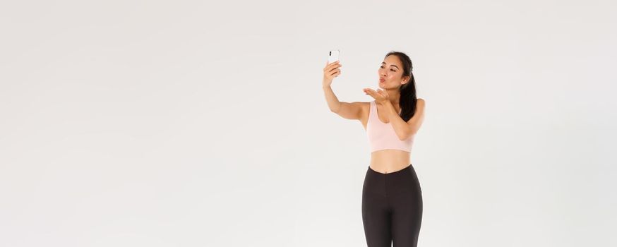 Full length of cute and silly female blogger in gym, wearing sportswear and taking selfie or live stream from gym workout session, sending air kiss at mobile phone screen, standing white background.