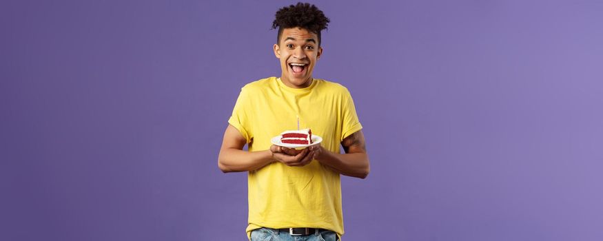 Happy birthday to me. Portrait of upbeat, excited hispanic man with dreads celebrating b-day, holding plate cake with lit candle, smiling amused, making wish, purple background.