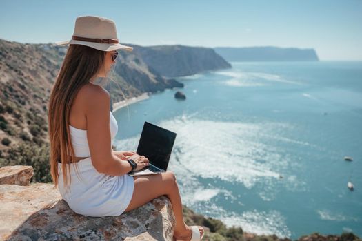 Successful business woman in yellow hat working on laptop by the sea. Pretty lady typing on computer at summer day outdoors. Freelance, travel and holidays concept.