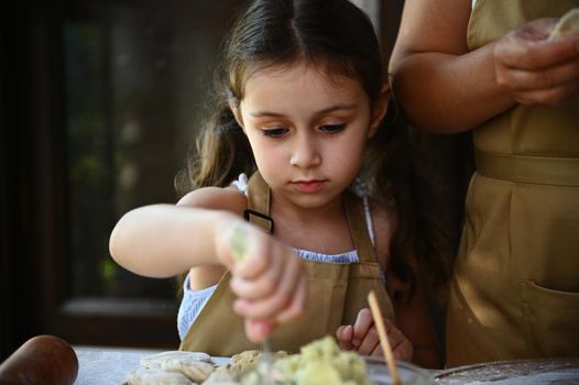 Adorable Caucasian little girl, child cook stuffing mashed potatoes into rolled out dough while learning cooking dumplings with her loving mother. Pretty girl preparing homemade Ukrainian vareniki