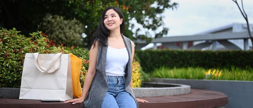 Happy asian woman with shopping bags sitting on a bench in the park.