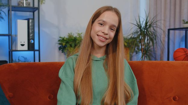 Close-up portrait of happy smiling teen girl in hoodie, looking at camera, celebrate good news. Young child kid indoor isolated at home in living room sitting on orange couch. Female nature beauty