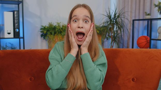 Oh my God, Wow. Lovely teen girl surprised looking at camera with big eyes, shocked by sudden victory, good win news, celebrating at home. Excited amazed young child kid indoor in living room on couch