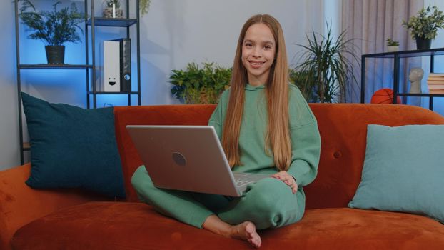 Lovely teen girl freelancer at home living room and sitting on orange couch sofa, opens laptop. Young child kid working on notebook, sends messages, makes online purchases, watching movies, working