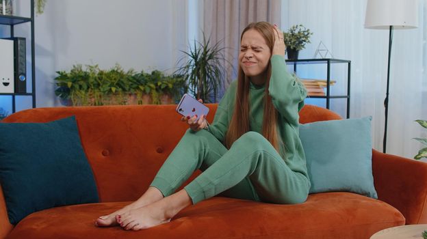 Portrait of caucasian girl use smartphone typing browsing, loses becoming surprised sudden lottery results, bad news, fortune loss, fail. Young child kid sitting on orange couch at home living room