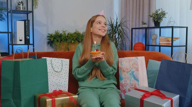 Happy teen girl wears festive birthday hat hold cupcake makes wish joyful congratulating blowing burning candle on cake. Young child kid celebrating anniversary party at home room apartment on couch