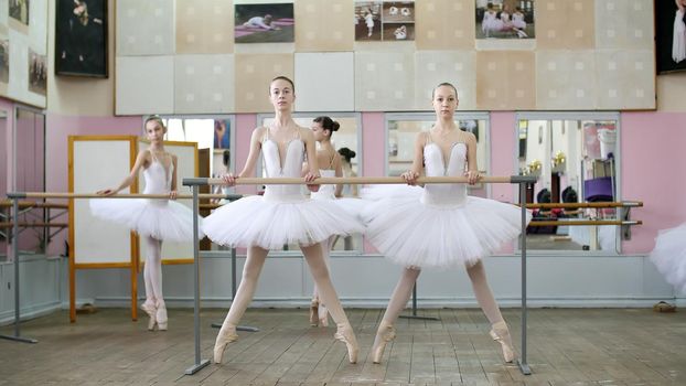 in the ballet hall, girls in white ballet tutus, packs are engaged at ballet, rehearse plie forward, Young ballerinas crouch standing on toes in pointe shoes at railing in ballet hall. High quality photo
