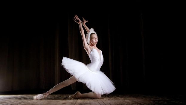 in rays of spotlight, on the stage of the old theater hall. Young ballerina in suit of white swan and pointe shoes, dances elegantly certain ballet motion, part de bras. High quality photo