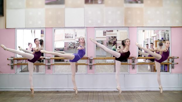 in dancing hall, Young ballerinas in purple leotards perform attitude efface on pointe shoes, standing near barre at mirror in ballet class. High quality photo