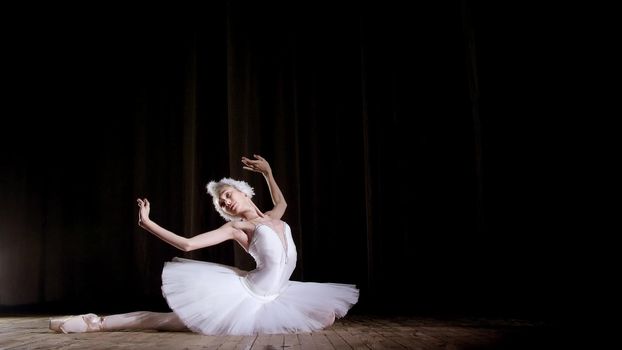 in rays of spotlight, on the stage of the old theater hall. Young ballerina in suit of white swan and pointe shoes, dances elegantly certain ballet motion, Swan Lake. High quality photo