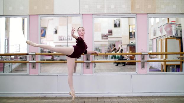 in dancing hall, Young ballerina in purple leotard performs developpe attitude on pointe shoes, raises her leg up behind elegantly, standing near barre at mirror in ballet class. High quality photo