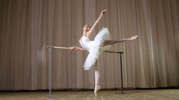 ballet rehearsal, in old theater hall. Young ballerina in white ballet skirt, tutu, is engaged at ballet, performs elegantly a certain ballet exercise, attitude allonge, standing near barre. High quality photo