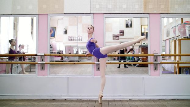 in dancing hall, Young ballerina in purple leotard performs developpe attitude on pointe shoes, raises her leg up behind elegantly, standing near barre at mirror in ballet class. High quality photo