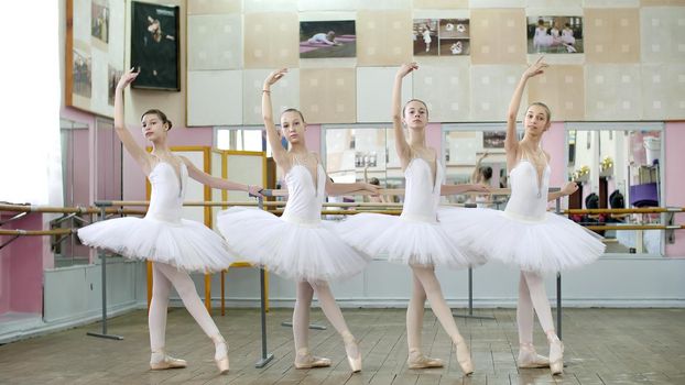 in ballet hall, girls in white ballet skirts are engaged at ballet, rehearse tendue forward battement, Young ballerinas standing in pointe shoes, at railing in ballet hall. High quality photo