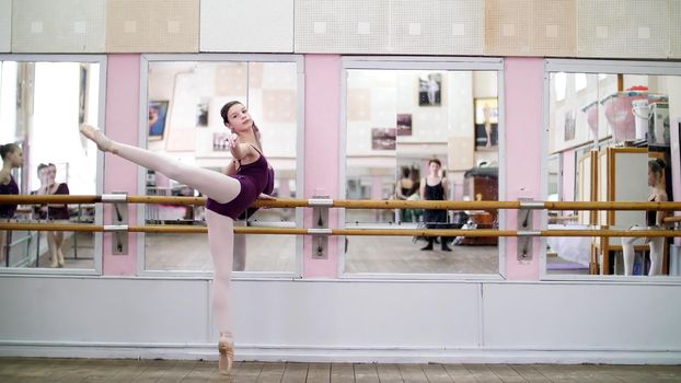 in dancing hall, Young ballerina in purple leotard performs developpe attitude on pointe shoes, raises her leg up behind elegantly, standing near barre at mirror in ballet class. High quality photo