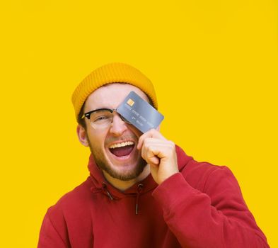 Young guy with credit or debit card in hand happy smiling cover his eye ready to spend money. Young man in red hoodie with mockup bank card isolated on yellow background. Shopping concept.