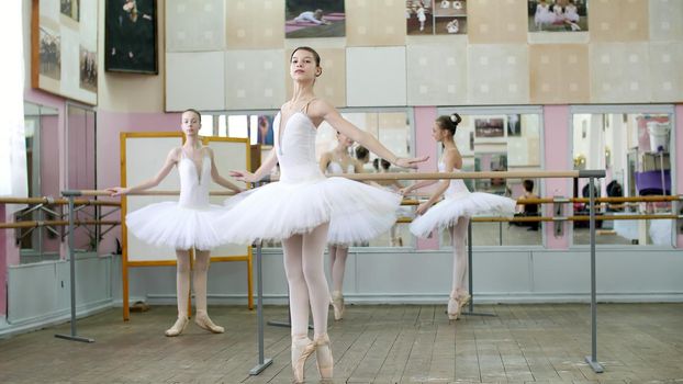 in ballet hall, girls in white ballet skirts are engaged at ballet, rehearse turning, Young ballerinas standing on toes in pointe shoes elegantly, at railing in ballet hall. High quality photo