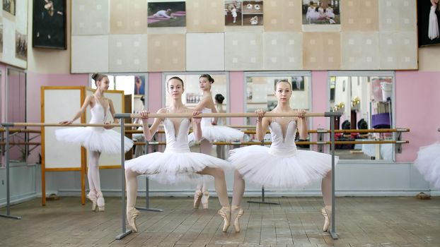 in the ballet hall, girls in white ballet tutus, packs are engaged at ballet, rehearse plie forward, Young ballerinas crouch standing on toes in pointe shoes at railing in ballet hall. High quality photo