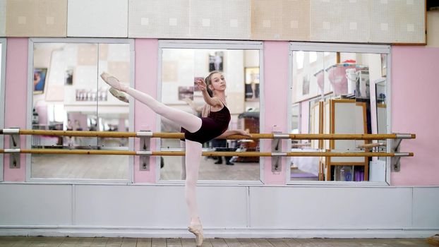 in dancing hall, Young ballerina in purple leotard performs developpe attitude on pointe shoes, raises her leg up behind elegantly, standing near barre at mirror in ballet class. High quality photo