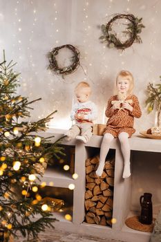 Little brother and sister play on Christmas eve in a beautiful house decorated for the New Year holidays. Children are playing with a Christmas gift. Scandinavian-style interior with live fir trees and a wooden staircase.