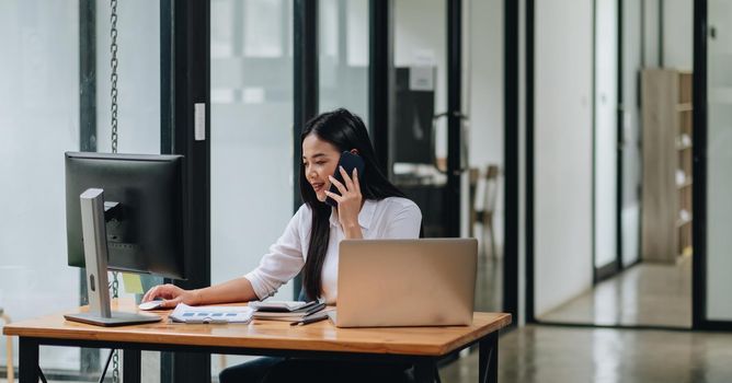 Image of asian woman talking on smartphone and working on laptop, while sitting at table indoor. business financial concept.