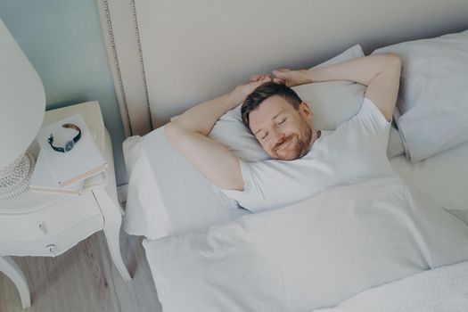 View from above of young happy relaxed caucasian man with attractive smile sleeping, lying in cozy bed with white beddings. Healthy care good night sleep and rest concept