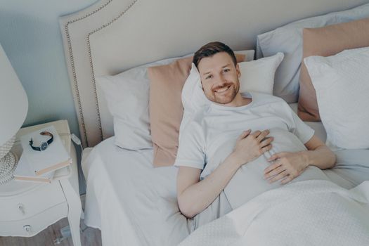 Young well rested man in white tee shirt waking up in morning with smile, happy after sleeping enough, lying in comfortable bed while covered with blankets, lots of different pillows