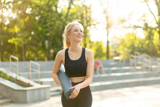 Sporty Slim Woman Sportive Clothes Black Leggings and Top Standing near concrete Stairs Holding Yoga Mat Resting Between Exercises. Sport and Fitness Concept Caucasian Athletic Female goes Workout