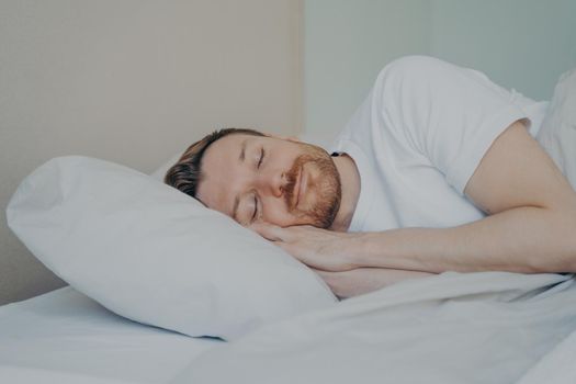Close up view photo of bearded young male with eyes closed fall asleep on his vacation on sunday, sleeping with smile while lying on white pillow in bedroom. Sweet dreaming and rest concept