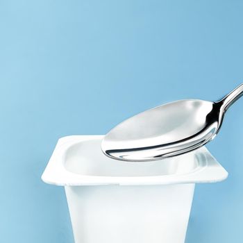 Yogurt cup and silver spoon on blue background, white plastic container, fresh dairy product for healthy diet and nutrition.