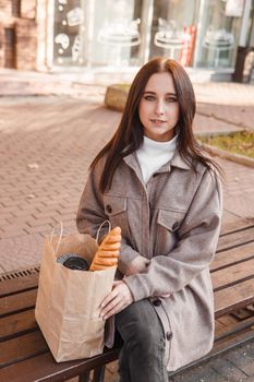 A stylish brunette woman walks around the autumn city. The brunette is sitting on a bench outside with a glass of coffee and a phone, holding a craft bag with a baguette.